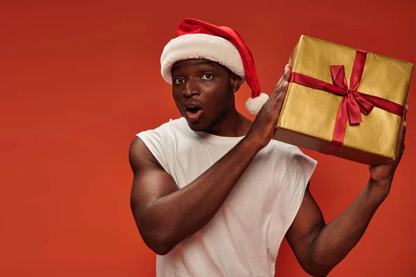 Sorprendido afroamericano hombre en santa hat celebración caja de regalo y mirando a la cámara en el fondo rojo - foto de stock