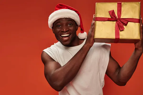 Bonhomme afro-américain ravi dans santa chapeau tenant boîte cadeau et regardant la caméra sur fond rouge — Photo de stock