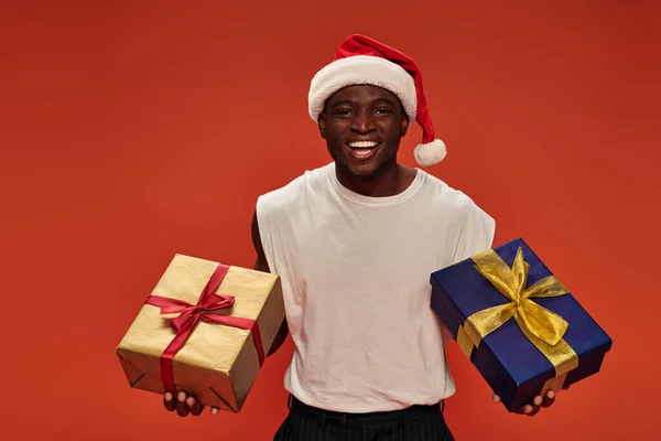 Радісний афроамериканський чоловік в різдвяному капелюсі і білий танк зверху позує зі святковими подарунками на червоному — стокове фото