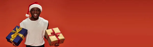 Delighted african american man in santa hat posing with colorful christmas presents on red, banner — Stock Photo