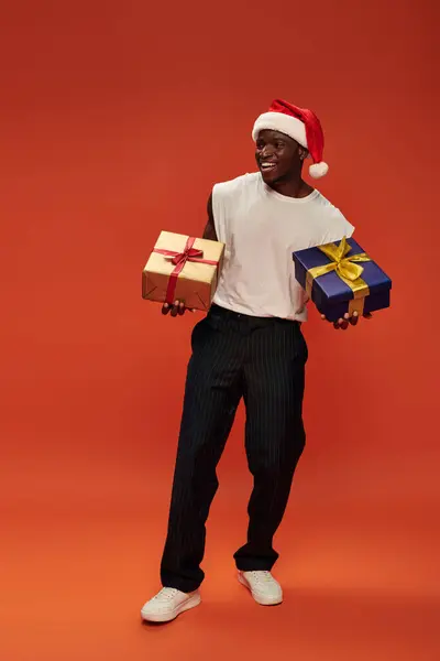 Повна довжина веселого афроамериканця в повсякденному одязі і шапці Санти, що тримає подарункові коробки на червоному — стокове фото