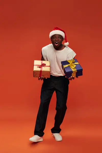 Emocionado hombre afroamericano en traje casual y sombrero de santa celebración de regalos de Navidad en rojo - foto de stock