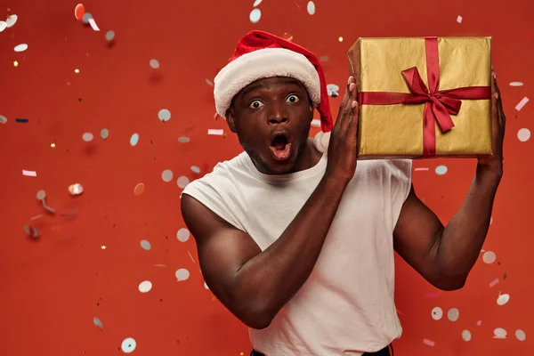 Amazed african american man in santa hat with presents and open mouth on red backdrop with confetti — Stock Photo