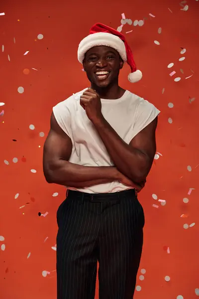 Joyful african american man in christmas hat looking at camera on red backdrop with falling confetti — Stock Photo