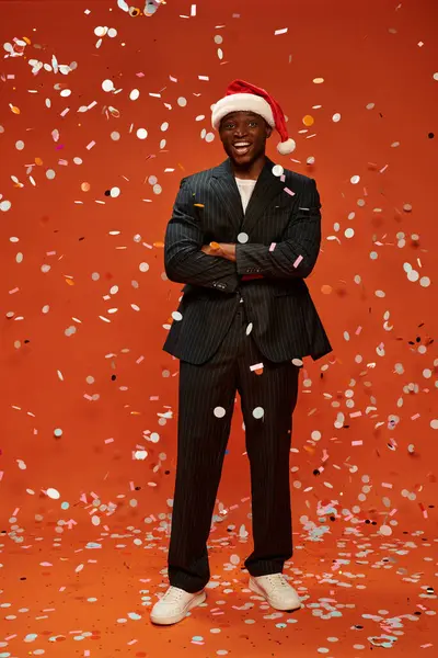 Hombre afroamericano excitado en traje negro y sombrero de santa sonriendo bajo confeti sobre fondo rojo - foto de stock