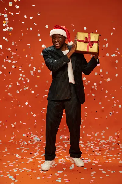Веселий афроамериканський чоловік в елегантному костюмі і капелюсі Санти тримає подарунок під конфетті на червоному — стокове фото