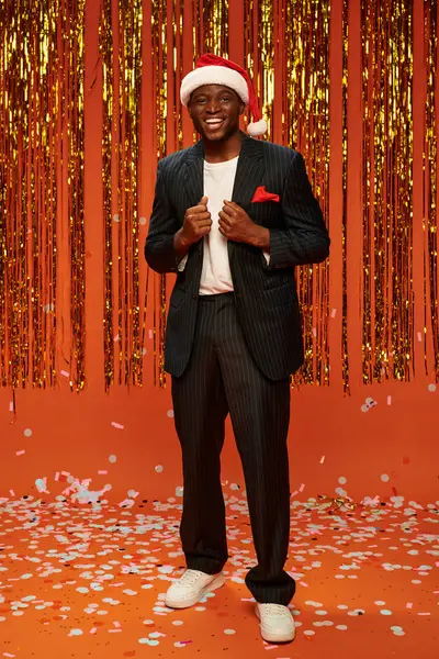 Cheerful african american man in santa hat and black suit on orange backdrop with shiny decor — Stock Photo