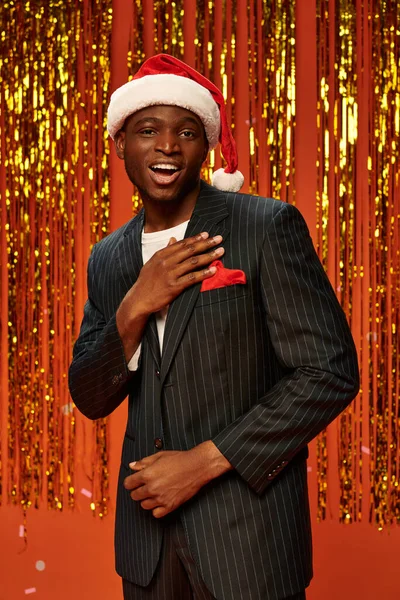 Кокетливый африканский американец в элегантном костюме и кепке Санты, смотрящий в камеру рядом с золотой мишурой — стоковое фото