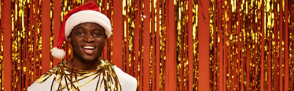 Веселий афроамериканський чоловік у шапці Санта, посміхаючись на камеру біля блискучої мішури на червоному, банер — стокове фото