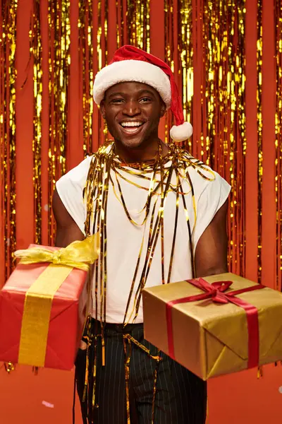 Счастливый африканский американец в шапочке Санты с рождественскими подарками рядом с золотой мишурой на красном — стоковое фото