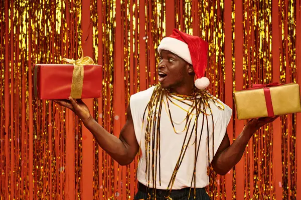 Asombrado hombre afroamericano en sombrero de santa con regalos de Navidad cerca de oropel brillante sobre fondo rojo - foto de stock