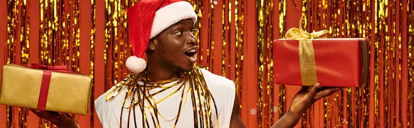 Erstaunt afrikanisch-amerikanischer Mann in Weihnachtsmütze mit Geschenkschachteln auf rotem Hintergrund mit glänzendem Lametta, Banner — Stockfoto