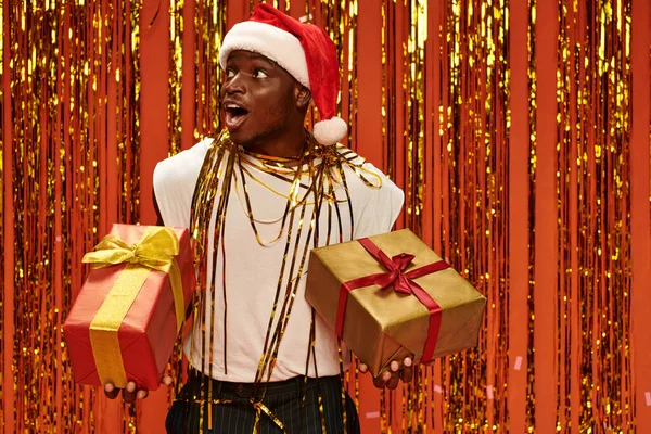 Дивовижний афроамериканський чоловік у шапці Санта тримає подарунки і дивиться далеко біля золотої блискучої мішури — стокове фото