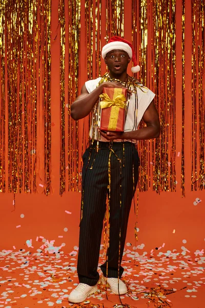 Здивований афроамериканський чоловік у шапці Санта, тримає подарунки і дивиться на камеру біля блискучого декору — стокове фото