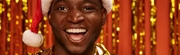 Portrait of laughing african american guy in santa near shiny golden tinsel on red backdrop, banner — Stock Photo