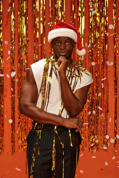 Riflessivo uomo africano americano in cappello di Babbo Natale e abiti casual vicino fili di lame d'oro su sfondo rosso — Foto stock