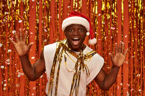 Amazed african american guy in santa hat and white tank top showing wow gesture on shiny backdrop — Stock Photo