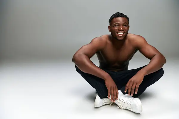 Cheerful african american guy with strong shirtless body smiling while sitting on grey, full length — Stock Photo
