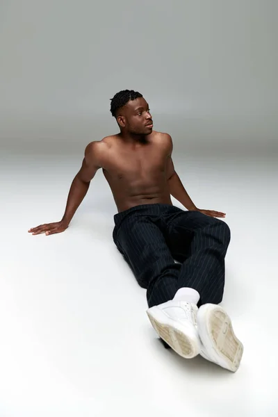 Full length of shirtless and athletic african american male model sitting and looking away on grey — Stock Photo