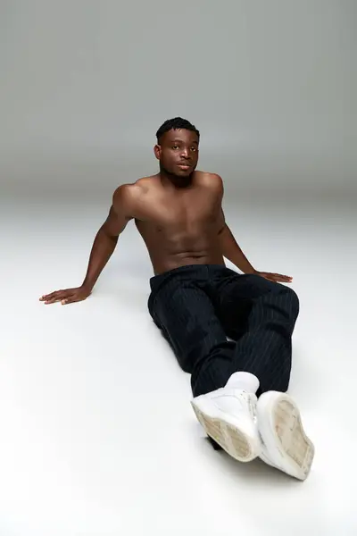 Volle Länge der hemdlosen und muskulösen afrikanisch-amerikanischen Mann sitzt und schaut in die Kamera auf grau — Stockfoto