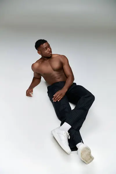 Confident shirtless african american fitness model in black pants sitting and looking away on grey — Stock Photo