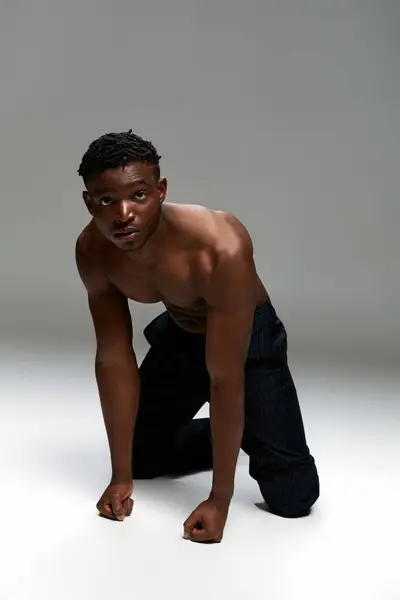 Muskulöser afrikanisch-amerikanischer Mann in schwarzer Hose auf Knien stehend und in die Kamera blickend auf grau — Stockfoto