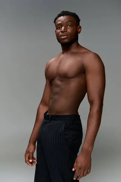 Американская модель фитнеса без рубашки в черных штанах, стоящая и смотрящая в камеру на сером — стоковое фото