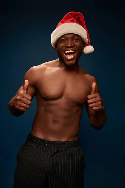 Eccitato e senza maglietta africano americano uomo in cappello di Babbo Natale mostrando pollici su sfondo blu navy — Foto stock