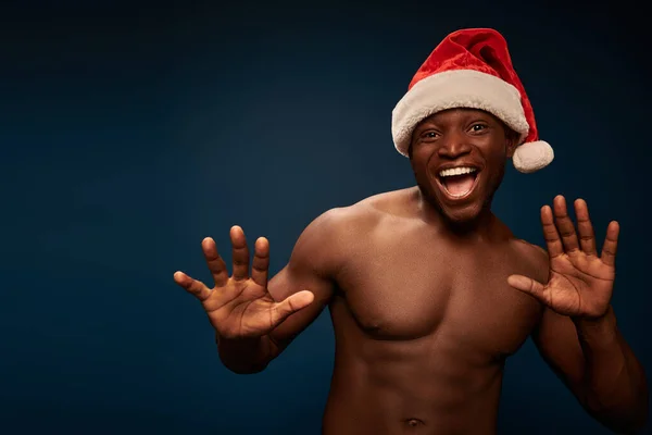 Überglücklicher afrikanisch-amerikanischer Mann mit muskulösem Oberkörper und Weihnachtsmütze gestikuliert vor dunkelblauem Hintergrund — Stockfoto