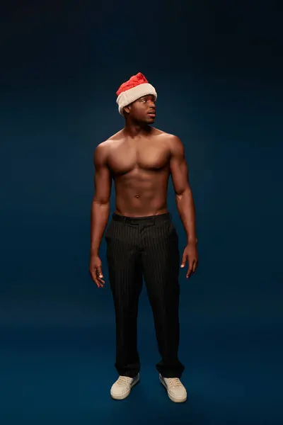 Muscular african american man in santa hat and black pants looking away in dark blue studio — Stock Photo
