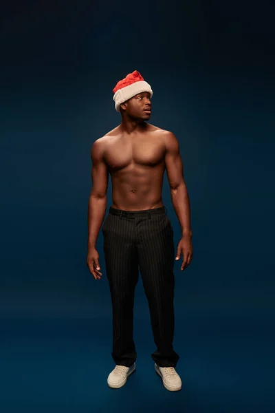 Muscular african american man in santa hat and black pants looking away in dark blue studio — Stock Photo