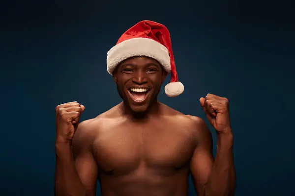 Joyful african american man with muscular torso wearing santa hat and rejoicing on dark blue — Stock Photo