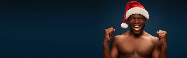 Joyeux sportif afro-américain en santa chapeau montrant geste de victoire sur fond bleu marine, bannière — Photo de stock