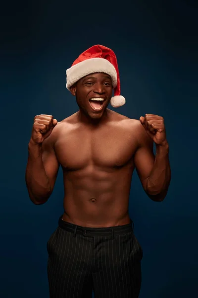 Overjoyed athletic african american in santa cap showing win gesture on navy blue backdrop — Stock Photo