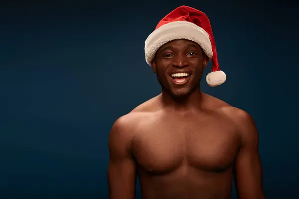 Glücklich und hemdlos afrikanisch-amerikanischer Kerl mit Weihnachtsmütze lächelt in die Kamera auf marineblauem Hintergrund — Stockfoto