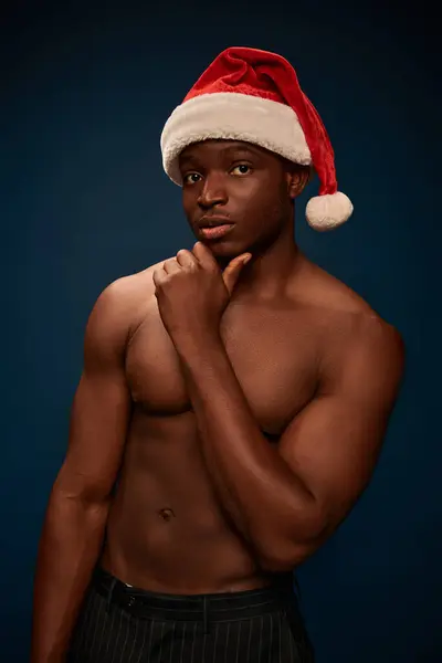 Thoughtful african american man in santa hat posing with shirtless torso on dark blue backdrop — Stock Photo