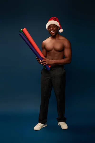 Cheerful muscular african american man in santa hat with colorful wallpapers on navy blue backdrop — Stock Photo