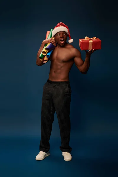 Shirtless and amazed african american man in santa hat with wallpapers and present on dark blue — Stock Photo