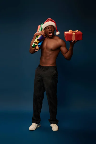 Sin camisa y sorprendido hombre afroamericano en sombrero de santa con fondos de pantalla y presente en azul oscuro - foto de stock