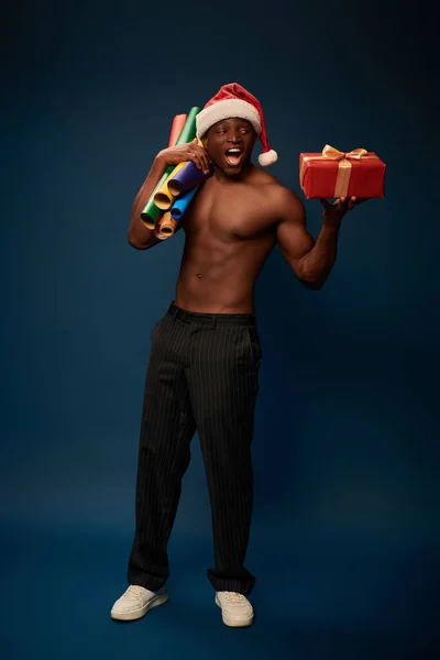 Thrilled and sportive african american man in santa hat with gift box and paper rolls on dark blue — Stock Photo
