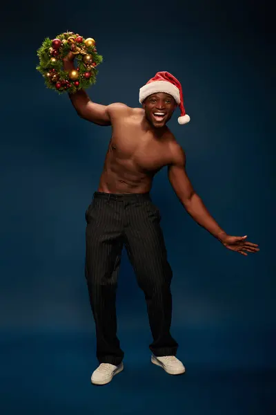 Fröhlich und hemdlos afrikanisch-amerikanischer Mann mit Weihnachtsmütze posiert Weihnachtskranz auf marineblau — Stockfoto