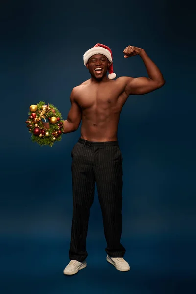 Astonished and athletic african american man in santa hat showing christmas wreaths on navy blue — Stock Photo
