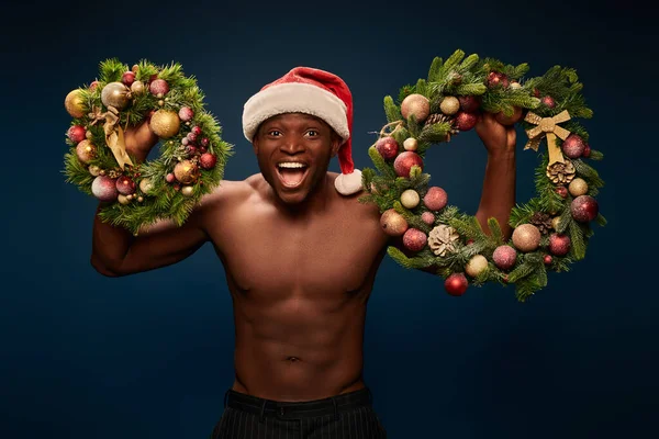 Без сорочки енергійний афроамериканський чоловік у капелюсі Санта, що показує різдвяні вінки на синьому — стокове фото
