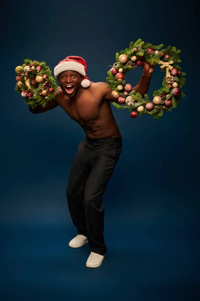 Impressed and muscular african american man in santa cap holding christmas wreaths on navy blue — Stock Photo