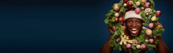 Portrait of cheerful american man in santa hat and christmas wreath on dark blue backdrop, banner — Stock Photo