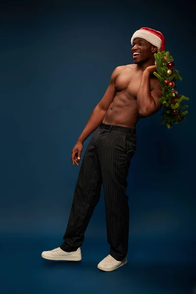 Glücklich athletisch afrikanisch-amerikanischer Mann mit Weihnachtsmütze mit Weihnachtskranz auf dunkelblauem Hintergrund — Stockfoto
