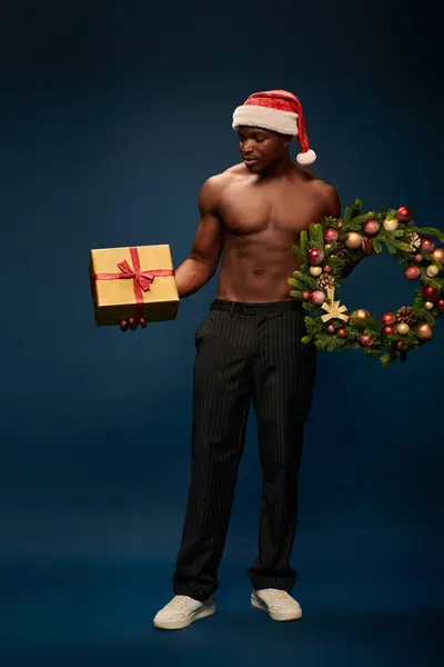 Hombre afroamericano sin camisa en santa hat celebración de caja de regalo y corona de Navidad en azul oscuro - foto de stock