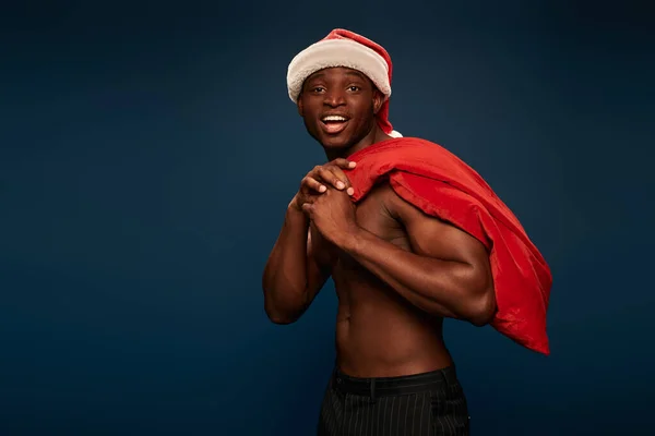 Animado e forte homem afro-americano em santa chapéu segurando saco de natal e sorrindo no azul escuro — Fotografia de Stock
