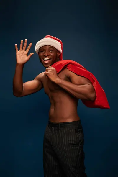 Hombre afroamericano feliz y sin camisa con bolsa de Navidad agitando la mano sobre fondo azul oscuro - foto de stock