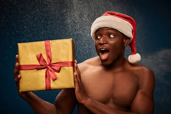 Изумленный безрубашечный африканский американец смотрит на блестящую подарочную коробку на синем снежном фоне — стоковое фото